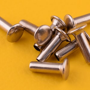 May include: A close-up of a pile of silver metal rivets on a yellow surface. The rivets have a round head and a long, thin shaft.