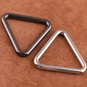 Op de afbeelding: Twee driehoekige metalen ringen, een zwart en een zilver, op een bruin suède oppervlak.
