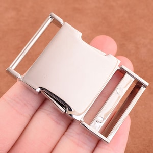 Puede incluir: Hebilla de metal plateado con una superficie lisa y plana y un mecanismo de cierre. La hebilla está diseñada para usarse con correas o cinturones.