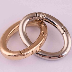 May include: Two oval-shaped metal clasps, one gold and one silver, are linked together. The clasps have a spring-loaded mechanism for opening and closing.