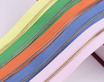 Puede incluir: Cinco cintas de cremallera de diferentes colores con dientes metálicos dorados y plateados. Las cintas están dispuestas en diagonal, con el color más claro en la parte superior y el color más oscuro en la parte inferior.