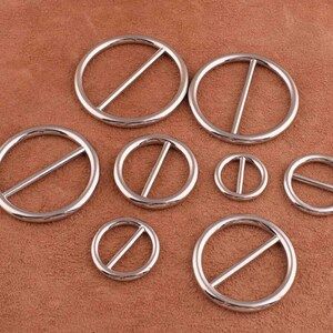 Puede incluir: Un conjunto de ocho anillos de metal plateado con una barra central. Los anillos son de diferentes tama&ntilde;os y est&aacute;n dispuestos en semic&iacute;rculo sobre una superficie de cuero marr&oacute;n.