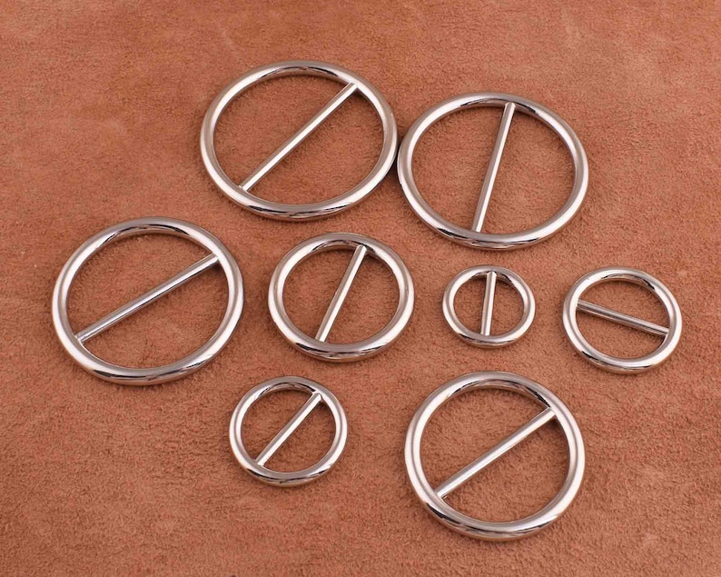 Puede incluir: Un conjunto de ocho anillos de metal plateado con una barra central. Los anillos son de diferentes tama&ntilde;os y est&aacute;n dispuestos en semic&iacute;rculo sobre una superficie de cuero marr&oacute;n.