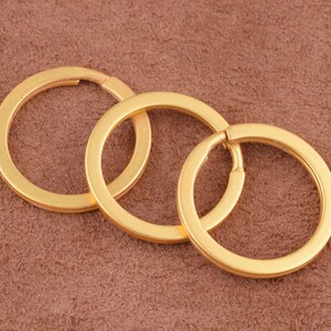 May include: Three gold-coloured metal key rings of different sizes, interlocked together. The key rings are on a brown suede-like surface.