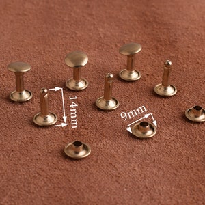 May include: A collection of antique brass rivets, each with a rounded head and a post for securing. The rivets are approximately 0.55 inches (14mm) in height and the base is 0.35 inches (9mm) in diameter. The rivets are displayed on a brown surface.