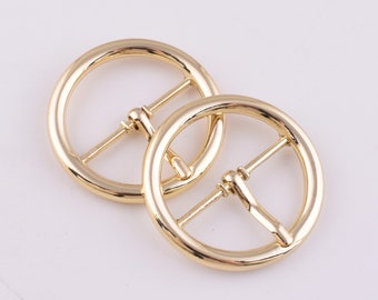May include: Two gold-coloured, circular belt buckles. Each buckle has a round frame with a central bar and a smaller bar. The buckles are made of shiny metal against a white background.