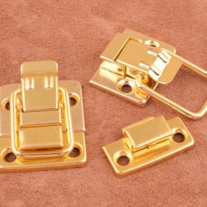 Könnte beinhalten: Drei goldfarbene Metallverschlüsse, zwei mit quadratischen und einer mit rechteckiger Basis. Die Verschlüsse haben einen Verriegelungsmechanismus und sind zum Sichern von Boxen, Taschen oder anderen Gegenständen konzipiert.