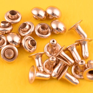 May include: A collection of 18 gold-toned metal rivets, some with a domed top and others with a flat top and a pointed post. The rivets are arranged on a yellow surface.