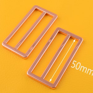 Puede incluir: Dos hebillas deslizantes de metal color oro rosa, cada una con tres barras horizontales. La hebilla de la derecha tiene 50 mm de ancho.