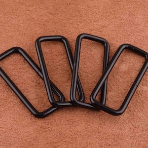 May include: Four black metal D-rings, each with a rectangular shape, are arranged in a loose pile on a brown textured surface.
