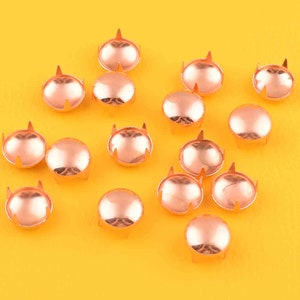 May include: A collection of 14 copper-colored metal studs with a rounded top and four prongs on the bottom. The studs are arranged on a yellow surface.