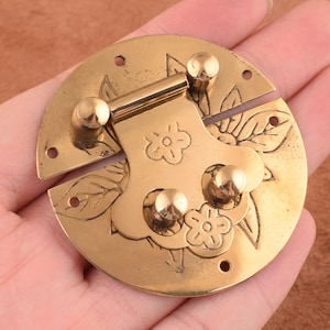 May include: A gold-toned metal latch with a floral design. The latch has two parts that open and close. The latch is designed to be used on a box, chest, or other piece of furniture.