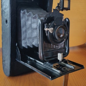 May include: A vintage black folding camera with a bellows system and a viewfinder. The camera has a lens with a silver ring and a black leather exterior.