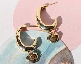 May include: Gold hoop earrings with a small heart charm dangling from each hoop. The heart charms have an eye design in the center.