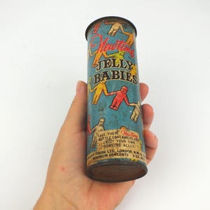 Könnte beinhalten: Eine zylindrische Vintage-Dose von Newtime Jelly Babies mit blauem Hintergrund und bunten Illustrationen von Gelee-Figuren. Der Text lautet Newtime JELLY BABIES, SAVE THESE LITTLE CONTAINERS AND ENJOY YOUR OWN BOWLING ALLEY, QUALITY FOODS LTD. LONDON, N.W.10, ENGLAND, MINIMUM CONTENTS 6 OZ. NET.
