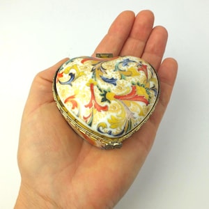Könnte beinhalten: Herzförmige Schmuckschatulle mit goldfarbenem Metallrahmen und floralem Design. Der Deckel hat einen cremefarbenen Hintergrund mit roten, gelben und blauen Blumenmustern. Die Schatulle wird in einer Hand gehalten.