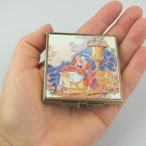 Könnte beinhalten: Ein quadratischer, vintage Kompakt-Make-up-Spiegel mit einem goldfarbenen Metallrahmen. Der Deckel zeigt eine farbenfrohe Illustration von zwei Figuren in historischer Kleidung, einem Hund und einer großen Blumenvase. Der Hintergrund umfasst eine Landschaft mit lila und rosa Farbtönen.