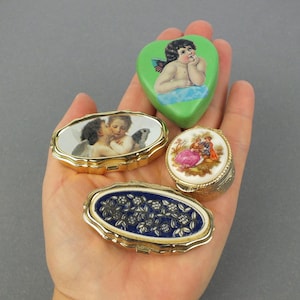 Op de afbeelding: Een verzameling vintage sieradendoosjes in verschillende vormen, waaronder ovaal en hartvormig. De doosjes hebben gedetailleerde kunstwerken, waaronder cherubs, bloemmotieven en romantische sc&egrave;nes. Goudkleurige metalen frames omlijsten de doosjes.