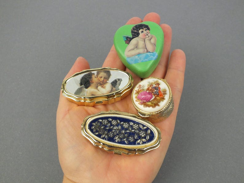 Op de afbeelding: Een verzameling vintage sieradendoosjes in verschillende vormen, waaronder ovaal en hartvormig. De doosjes hebben gedetailleerde kunstwerken, waaronder cherubs, bloemmotieven en romantische sc&egrave;nes. Goudkleurige metalen frames omlijsten de doosjes.