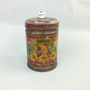 Könnte beinhalten: Vintage Kaffee-Blechdose mit Deckel und weißem Knauf. Die Dose hat ein farbenfrohes Design mit dem Wort "KAFFEE" und einer Illustration einer Frau, die von Blumen umgeben ist. Die Dose hat ein rot-, grün- und goldfarbenes Farbschema.