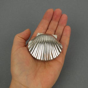 Könnte beinhalten: Ein silberfarbenes Metall-Kompaktgehäuse in Form einer Jakobsmuschel. Die Außenseite weist eine gerippte, fächerartige Textur mit metallischem Glanz auf. Das Objekt ist an der Oberseite mit einem Scharnier versehen und hat eine glatte Oberfläche.
