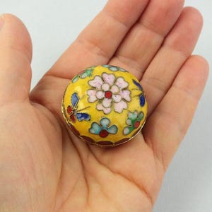 Könnte beinhalten: Kleine, runde, gelbe Cloisonné-Schmuckdose mit floralem Design. Die Dose hat eine zentrale rosa Blume mit roten und weißen Akzenten, umgeben von blauen und grünen Blumen- und Schmetterlingsmotiven. Sie hat einen goldfarbenen Metallrand.