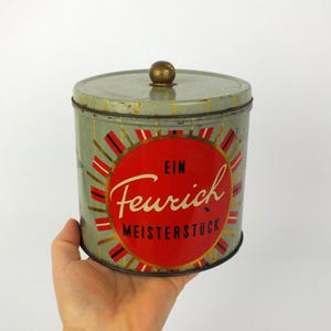 Könnte beinhalten: Vintage zylindrische Dose mit Deckel und Messingknopf. Die Dose ist hellgrün mit einem roten und weißen kreisförmigen Design in der Mitte, mit dem Wort "Feurich" in Schreibschrift. Die Wörter "EIN" und "MEISTERSTUCK" sind ebenfalls sichtbar.