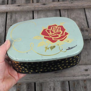Könnte beinhalten: Vintage ovale Blechdose mit hellgrünem Deckel, verziert mit einer roten Rose und goldenen Akzenten. Die Dose hat einen schwarzen Boden mit goldenen Punkten. Ein dekorativer Aufbewahrungsbehälter.