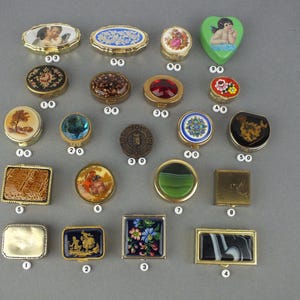 Op de afbeelding: Een verzameling kleine, decoratieve doosjes in verschillende vormen, waaronder ovaal, rond en vierkant. De doosjes hebben ingewikkelde ontwerpen, zoals cherubs, bloemmotieven en abstracte kunst. Het kleurenpalet omvat goud, blauw, groen en rood.
