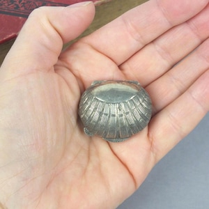 May include: A small, silver-coloured, shell-shaped trinket box. The box is held in an open hand, showcasing its detailed, ridged design. The box is round and has a hinged lid. The background includes a portion of a red book.