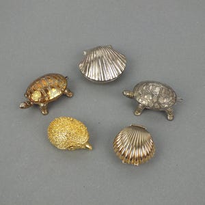 Könnte beinhalten: Eine Sammlung von fünf kleinen Schmuckdosen aus Metall, bestehend aus zwei schildkrötenförmigen Dosen in Bronze- und Silbertönen, zwei muschelförmigen Dosen in Silber- und Goldtönen sowie einer goldfarbenen igelförmigen Dose mit strukturierter Oberfläche.