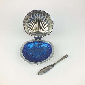 Könnte beinhalten: Eine silberfarbene, muschelförmige Schale mit Klappdeckel und einem leuchtend blauen Innenraum. Ein kleines, silberfarbenes Messer liegt daneben. Die Schale dient wahrscheinlich zum Servieren von Butter oder Gewürzen.