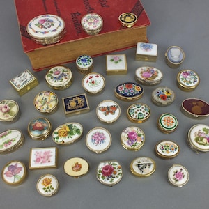 Könnte beinhalten: Eine Sammlung kleiner, dekorativer Schmuckdosen mit floralen und gemusterten Designs. Die Dosen haben verschiedene Formen, darunter rund, oval und rechteckig, mit goldfarbenen Metallrändern. Einige Dosen zeigen gemalte Szenen.