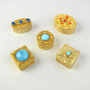Könnte beinhalten: Fünf verzierte, goldfarbene Schmuckkästchen mit verschiedenen Formen und Designs. Einige Kästchen weisen blaue oder rote Edelsteine auf, während andere aufwendige Filigran- oder Prägemuster haben. Die Kästchen sind auf einer weißen Oberfläche angeordnet.