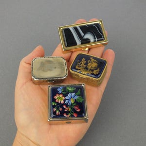 Op de afbeelding: Vier kleine, decoratieve doosjes met scharnierende deksels. E&eacute;n heeft een zwart-wit marmerpatroon, een ander heeft een blauw-gouden sc&egrave;ne, een derde heeft een bloemenpatroon en de laatste is zilverkleurig. De doosjes worden in een hand gehouden.