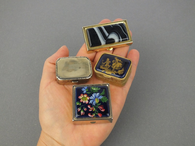 Op de afbeelding: Vier kleine, decoratieve doosjes met scharnierende deksels. E&eacute;n heeft een zwart-wit marmerpatroon, een ander heeft een blauw-gouden sc&egrave;ne, een derde heeft een bloemenpatroon en de laatste is zilverkleurig. De doosjes worden in een hand gehouden.