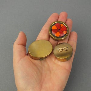 Könnte beinhalten: Drei kleine, messingfarbene Behälter. Ein runder Behälter hat eine Mosaikoberfläche mit roten und orangen Glasstücken. Ein weiterer runder Behälter hat eine Brille auf der Oberseite. Der dritte Behälter ist oval geformt.