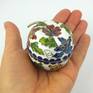 Könnte beinhalten: Eine weiße, apfelförmige Schmuckschatulle mit einem silbernen Stiel. Die Schatulle ist mit bunten Blumenmustern in Rot, Blau, Grün und Gelb verziert. Die Schatulle wird in einer Hand gehalten.