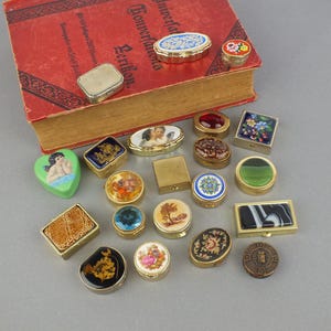 Op de afbeelding: Een verzameling decoratieve pillendoosjes in verschillende vormen en ontwerpen, waaronder ovaal, rond en rechthoekig. De doosjes hebben ingewikkelde patronen, bloemmotieven en miniatuurschilderijen. Sommige zijn goudkleurig, andere met kleurrijke emaille details.