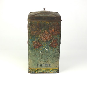 Könnte beinhalten: Eine antike Kaffeedose aus Metall mit einem Blumendesign und dem Wort "Kaffee" auf der Vorderseite. Die Dose ist rostig und hat eine verblichene grün-blaue Farbe.