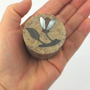 Könnte beinhalten: Kleine, runde, beige Steinbox mit floralem Design. Die Blume hat ein silbernes Blütenblatt und einen braunen Mittelteil, mit dunkelgrauen Blättern und Stiel. Die Box wird in einer Hand gehalten.