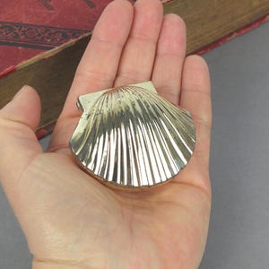 May include: A small, silver-coloured seashell-shaped trinket box. The box is held in an open hand, with the shell design clearly visible. The box has a ridged pattern radiating from the centre.