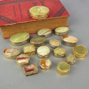 Könnte beinhalten: Eine Sammlung kleiner, dekorativer Schmuckdosen in verschiedenen Formen, darunter rund, oval und quadratisch. Die Dosen haben goldfarbene Metallränder und Deckel aus poliertem Stein in Grün-, Braun- und Cremetönen. Im Hintergrund liegt ein altes Buch.