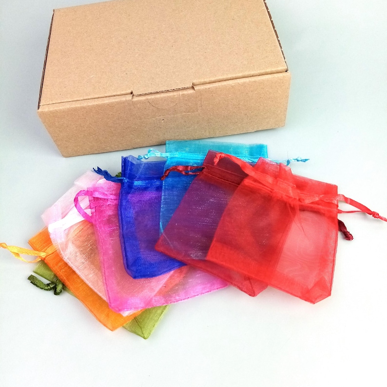 Op de afbeelding: Een verzameling kleurrijke, transparante organza cadeauzakjes in verschillende kleuren, waaronder oranje, roze, blauw en rood, zijn in een waaier gerangschikt. Een bruine kartonnen doos staat op de achtergrond. De zakjes hebben trekkoordsluitingen.