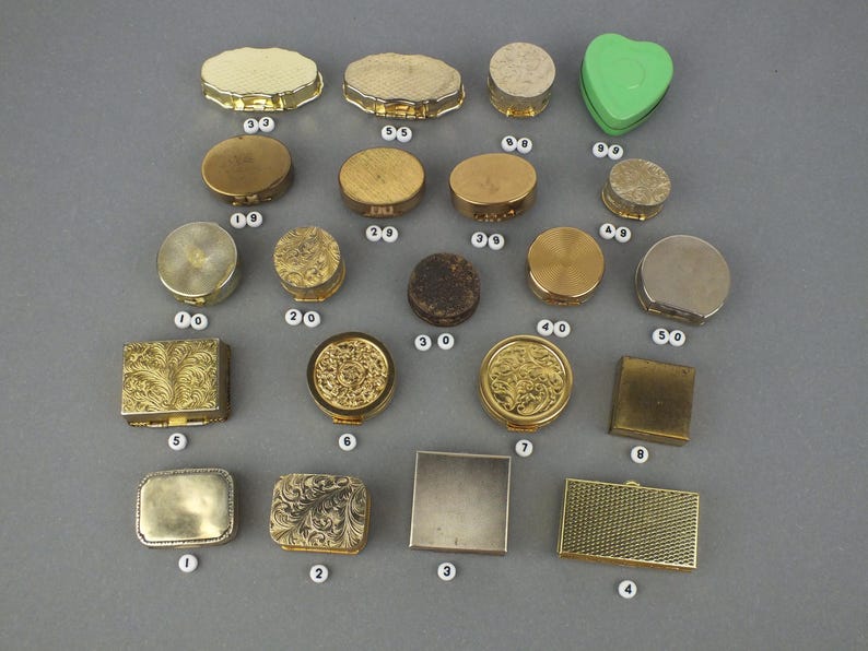 Op de afbeelding: Een assortiment vintage pillendoosjes in verschillende vormen en maten. De collectie omvat ronde, ovale en vierkante doosjes, sommige met sierlijke gouden details en andere met een eenvoudige, metalen afwerking. Er is ook een groen hartvormig doosje aanwezig.