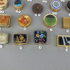 Op de afbeelding: Een verzameling kleine, decoratieve doosjes met verschillende ontwerpen. De doosjes zijn rond, vierkant en rechthoekig, met ingewikkelde patronen, bloemmotieven en kleurrijke accenten. Sommige hebben gouden of zilveren frames, andere een edelsteen of glazen bovenkant.