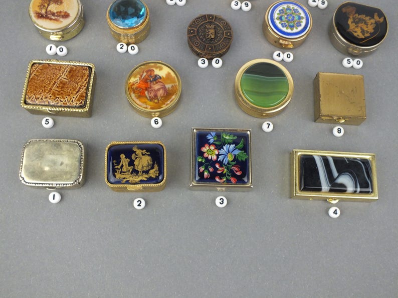 Op de afbeelding: Een verzameling kleine, decoratieve doosjes met verschillende ontwerpen. De doosjes zijn rond, vierkant en rechthoekig, met ingewikkelde patronen, bloemmotieven en kleurrijke accenten. Sommige hebben gouden of zilveren frames, andere een edelsteen of glazen bovenkant.