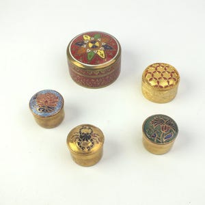 Könnte beinhalten: Fünf kleine, runde dekorative Schatullen mit aufwendigen Designs. Die Schatullen haben goldfarbene Metallbasen und farbenfrohe Emailledeckel mit floralen und geometrischen Mustern. Die größte Schatulle ist rot mit goldenem Rand.