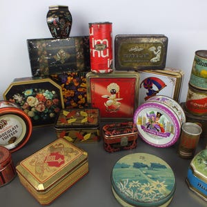 May include: A collection of various vintage metal storage tins in assorted shapes, sizes, and colours. The containers feature diverse graphic designs, including floral patterns, brand logos like Quality Street and Chocolate Toffees, and landscape illustrations. These decorative boxes are made of tin and display a range of colourful, weathered, and printed surfaces.
