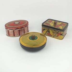 Op de afbeelding: Drie vintage metalen blikken. Een ovaal blik met een rood en wit ontwerp, een rechthoekig blik met een gouden achtergrond en bladerenpatroon, en een rond blik met een gouden deksel en zwarte basis.
